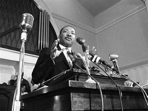 Black-and-white photo of Dr. Martin Luther King Jr. speaking at a church podium in Atlanta, surrounded by microphones, with his hands raised mid-gesture.