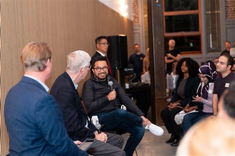 A panel discussion in a modern room, with one man speaking into a microphone while others listen. Audience members sit nearby, some smiling and engaged.