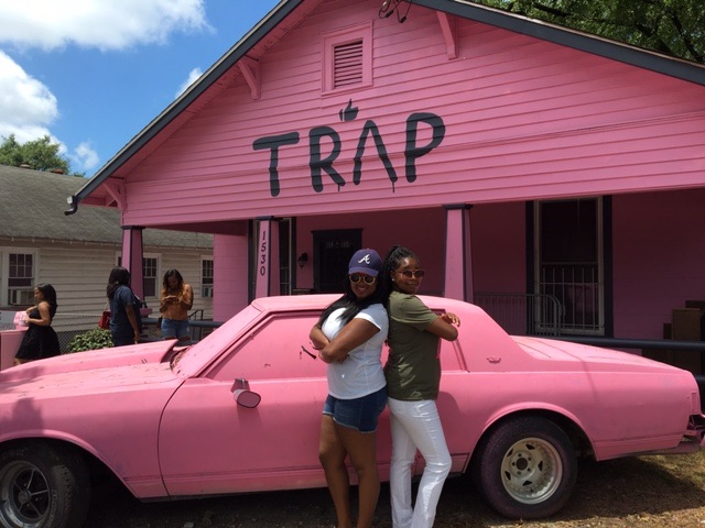Two people posing in front of a bright pink house with the word ‘TRAP’ stenciled in black across the front, with a matching pink car parked in front—2 Chainz’s famous Pink Trap House in Atlanta.
