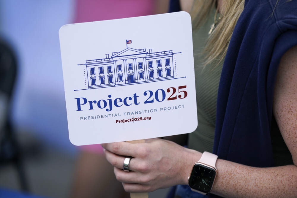 Close-up of a person holding a square sign that reads “Project 2025 Presidential Transition Project” with a line drawing of the White House above the text.