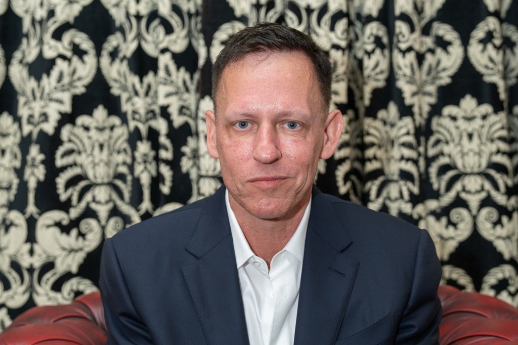 Close-up portrait of Peter Thiel in a navy suit and white shirt, seated against a patterned black-and-white backdrop, looking directly at the camera.