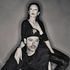 Black-and-white promotional photo of Phantogram. Sarah Barthel stands confidently behind Josh Carter, resting one hand on his shoulder. She wears a sleek black outfit, and he wears a dark shirt and a hat with the Phantogram “P” logo. The image has a moody, stylized tone.