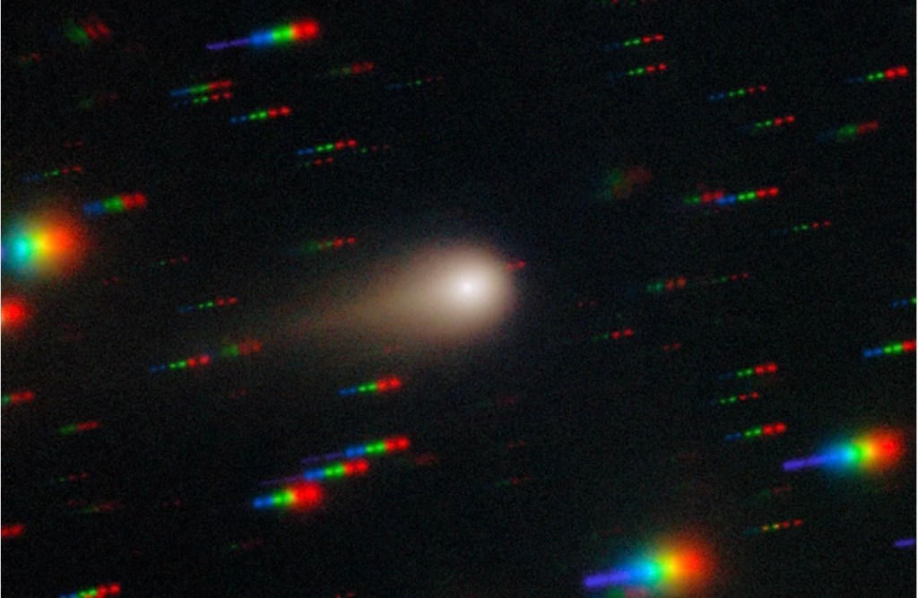 Real telescope image of interstellar comet 3I/ATLAS showing a glowing white nucleus and faint tail against a dark sky with multicolored star trails.