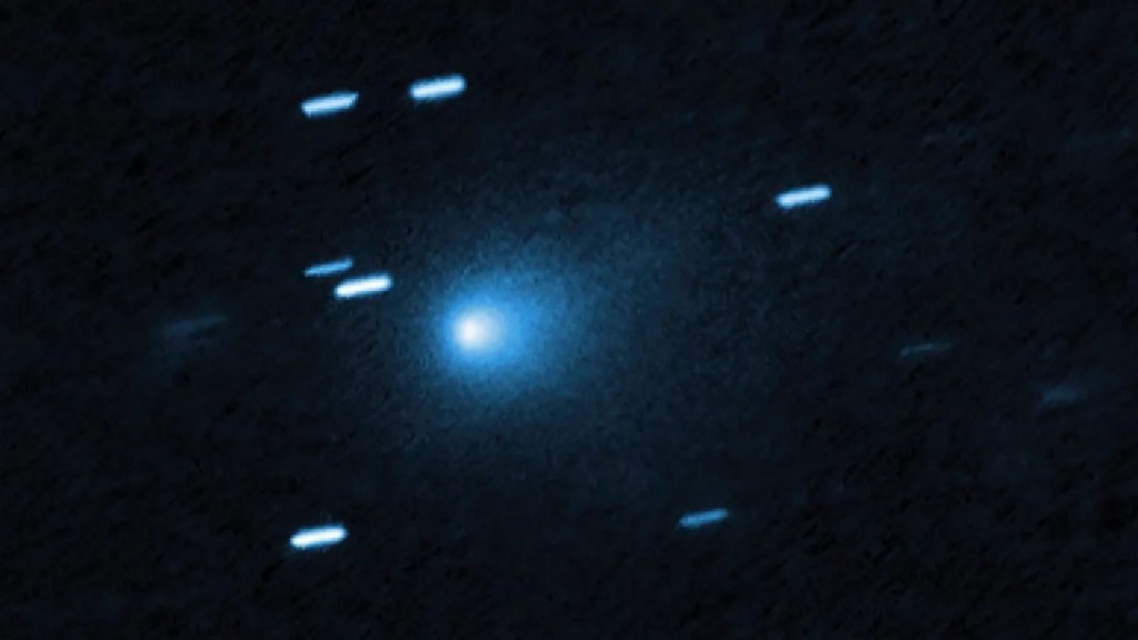 Real telescope image of interstellar comet 3I/ATLAS glowing bright blue at its core, surrounded by a faint halo of dust and gas, with background stars appearing as short blue streaks against a dark sky.